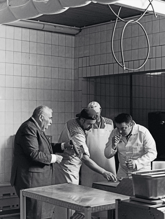 Das Bild zeigt eine Schwarz-Weiß-Fotografie von drei Männern in einer Küche, von denen einer einen Anzug trägt und die beiden anderen Schürzen. Sie scheinen sich zu unterhalten oder gemeinsam Essen zuzubereiten.