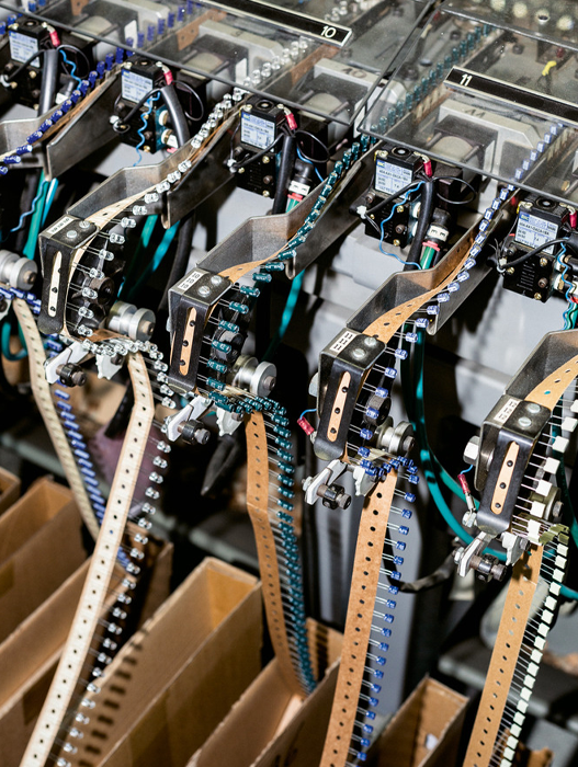 Das Bild zeigt eine Nahaufnahme einer komplexen Fertigungsstraße mit verschiedenen elektronischen Bauteilen und Maschinen. Die Bauteile sind durch ein Netz aus Drähten und Kabeln miteinander verbunden, wodurch eine optisch komplexe und detailreiche Szene entsteht. Das Bild wurde aus einem hohen Blickwinkel aufgenommen und bietet einen umfassenden Überblick über die Fertigungsstraße und ihre komplexen Details. Der Hintergrund ist unscharf, wodurch der Fokus auf die Maschinen und ihre Komponenten gelenkt wird. Das Bild enthält keinen erkennbaren Text, und die relative Anordnung der Objekte lässt auf eine gut organisierte und funktionale Fertigungsstraße schließen.