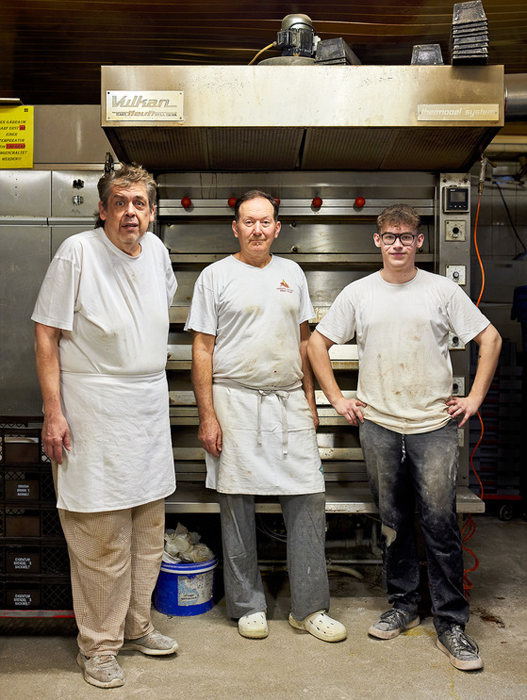 Drei Männer in weißen Schürzen stehen in einer Bäckerei vor einem großen Industrieofen. Der Mann links trägt eine Brille und scheint der Älteste der Gruppe zu sein. Der Mann in der Mitte trägt ein rotes Logo auf seinem Hemd. Der Mann rechts trägt eine Brille und wirkt eher leger. Der Ofen hinter ihnen ist ein markantes Merkmal der Bäckerei; darüber hängt ein Schild mit der Aufschrift „Milan“.