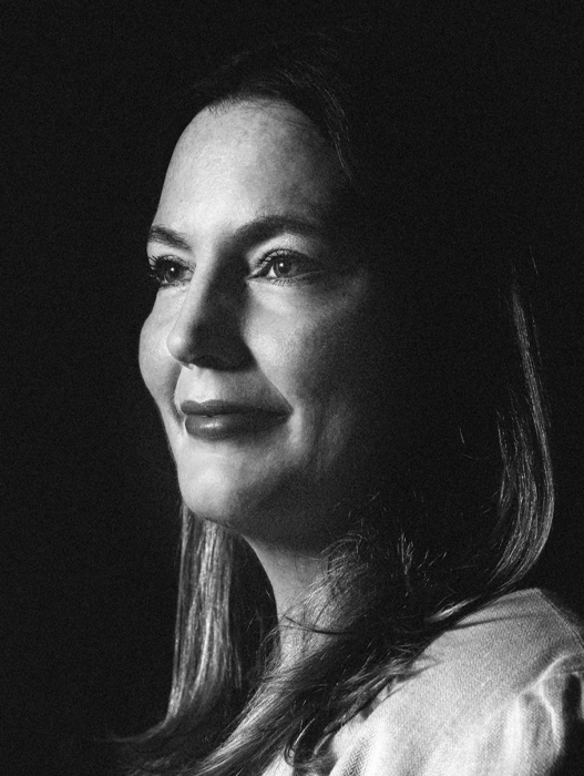Das Bild ist ein Schwarz-Weiß-Porträt einer Frau mit langen Haaren, aufgenommen in Nahaufnahme. Sie lächelt, ihr Blick ist zur Seite gerichtet, was ein Gefühl von Tiefe und Emotion vermittelt. Das Bild ist im Hochformat gehalten, wodurch die Gesichtszüge und der Ausdruck der Frau besonders hervorgehoben werden. Die Gesamtkomposition ist schlicht und fokussiert, wobei die Frau im Mittelpunkt steht.