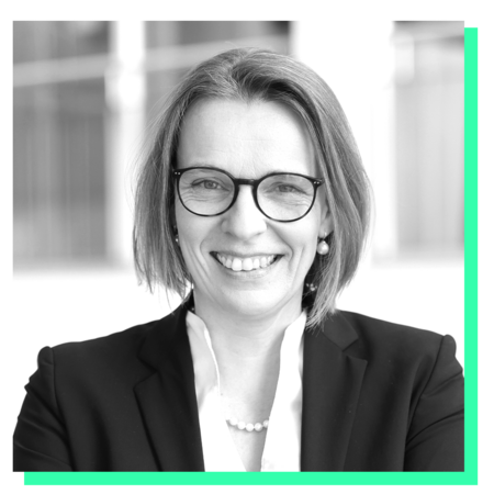 Das Bild zeigt ein Schwarz-Weiß-Porträt einer Frau mit kurzen Haaren und Brille, die in die Kamera lächelt. Sie trägt einen Blazer und eine Perlenkette um den Hals. Der Hintergrund ist unscharf, was auf eine Innenaufnahme hindeutet, möglicherweise einen Raum mit Fenstern. Das Bild ist von einem grünen Rahmen umgeben, was ihm ein professionelles und gepflegtes Aussehen verleiht.