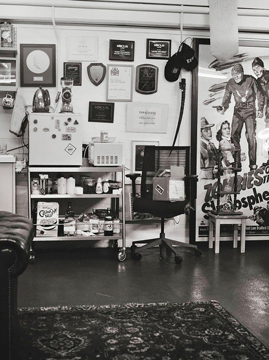 Das Bild zeigt eine Schwarz-Weiß-Fotografie eines überfüllten Raums mit verschiedenen Gegenständen, darunter ein Schreibtisch, ein Stuhl, ein Poster und ein Regal. Der Raum scheint ein Arbeits- oder Abstellraum zu sein, mit einem Teppich auf dem Boden und einem Fenster im Hintergrund. Die Gegenstände im Raum sind etwas ungeordnet angeordnet, was auf eine geschäftige oder kreative Umgebung hindeutet.