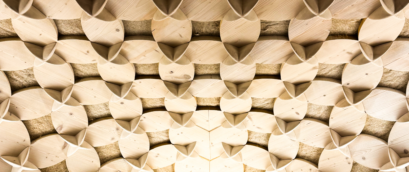 Das Bild zeigt eine Nahaufnahme einer Holzdecke mit einem sich wiederholenden Muster aus Holzpaneelen, die in einer rautenförmigen Anordnung angeordnet sind. Die Paneele sind hellbraun und weisen eine strukturierte Oberfläche auf, wodurch ein optisch ansprechendes und filigranes Muster entsteht. Das Bild enthält keinen erkennbaren Text und keine zählbaren Objekte, und die relative Anordnung der Paneele bleibt im gesamten Muster gleich, was zur Gesamtästhetik der Decke beiträgt.