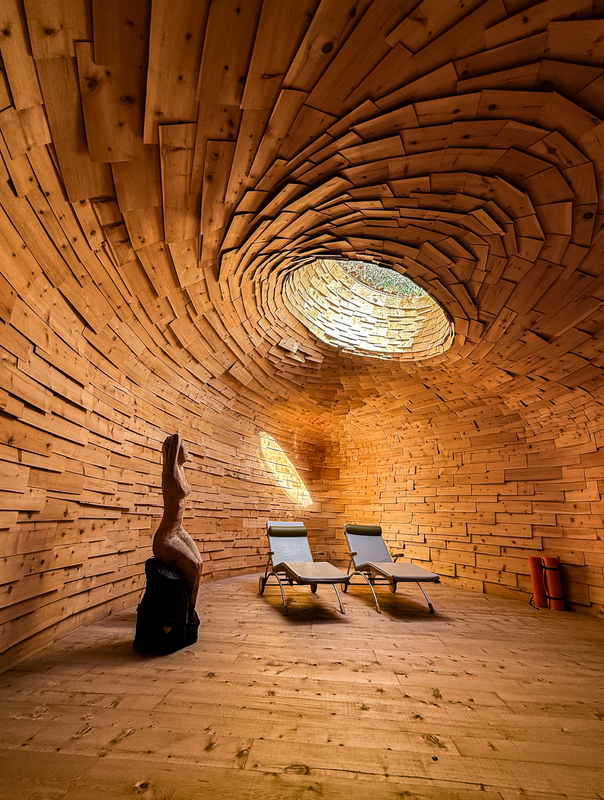 Das Bild zeigt einen ruhigen Innenraum mit Holzdecke und -wänden, der eine warme und einladende Atmosphäre schafft. In der Mitte des Raumes stehen zwei sich gegenüberliegende Sessel, die eine gemütliche Sitzecke bilden. Auf einem Hocker steht eine Skulptur einer menschlichen Figur, die dem Raum eine künstlerische Note verleiht. Der Raum wird durch Tageslicht erhellt, das durch ein rundes Fenster hereinströmt und das Gesamtambiente unterstreicht.