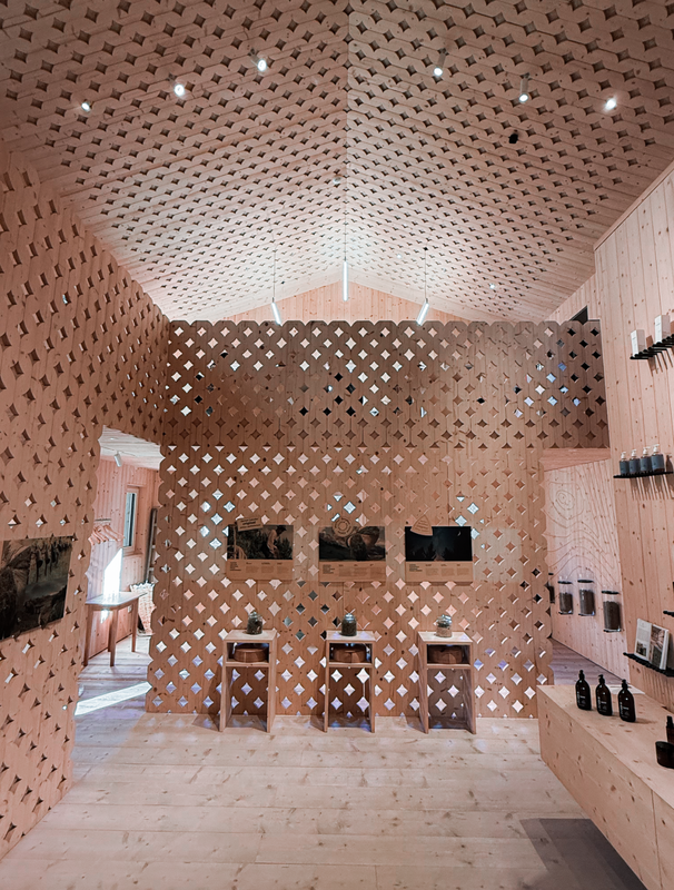 Das Bild zeigt einen modernen Innenraum mit einer Wand aus Holzgitter. Vor der Wand stehen drei Holzhocker, und auf einem Regal rechts sind mehrere Flaschen ausgestellt. Der Raum verfügt über eine hohe Decke mit Einbauleuchten, was eine helle und luftige Atmosphäre schafft. Das Gesamtdesign wirkt minimalistisch und funktional, wobei der Schwerpunkt auf der Verwendung natürlicher Materialien und offenen Flächen liegt.