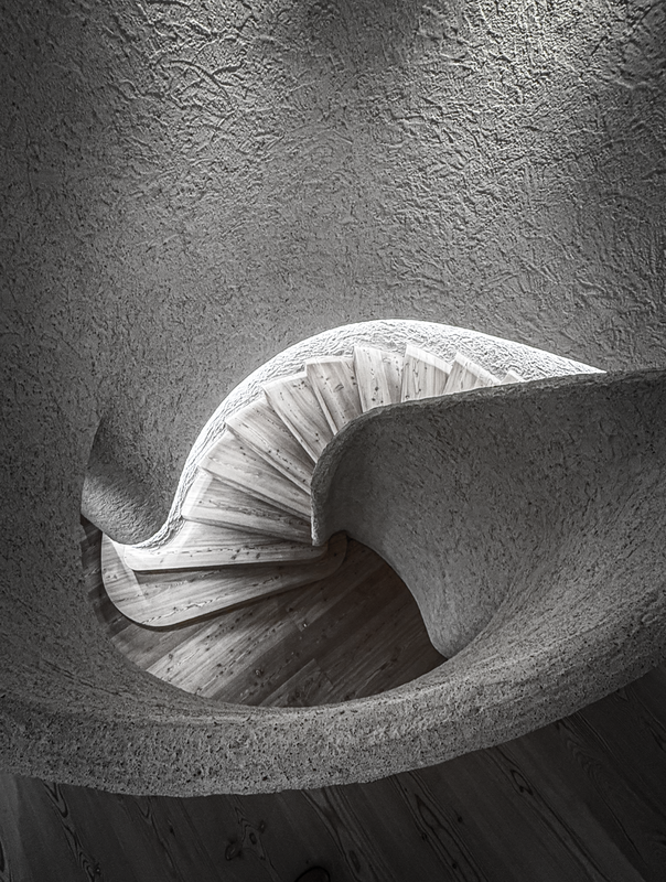 Das Bild zeigt eine Wendeltreppe mit Holzstufen und einer geschwungenen Betonwand. Die Treppe steht vor einer strukturierten Wand, wodurch ein visuell eindrucksvoller Kontrast entsteht. Das Bild ist in Schwarz-Weiß gehalten, was der Szene einen zeitlosen Charakter verleiht. Die Treppe scheint Teil einer größeren architektonischen Struktur zu sein, möglicherweise eines Gebäudes oder eines Museums, wobei die Holzstufen und die geschwungene Wand ein Gefühl von Tiefe und Dimension vermitteln. Das Bild ist gut ausbalanciert: Die Treppe befindet sich in der Mitte und die Wand auf der rechten Seite, wodurch eine harmonische Komposition entsteht.