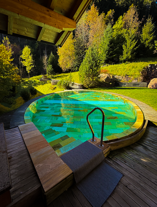 Das Bild zeigt eine ruhige Außenanlage mit einem runden Pool, der von einer Holzterrasse umgeben ist. Der Pool ist mit Wasser gefüllt und verfügt auf der rechten Seite über ein Metallgeländer. Die Terrasse besteht aus Holz und verfügt über einen Steinweg, der zum Pool führt. Die Szene ist von üppigem Grün umgeben, darunter Bäume und Sträucher, was eine ruhige und malerische Atmosphäre schafft.