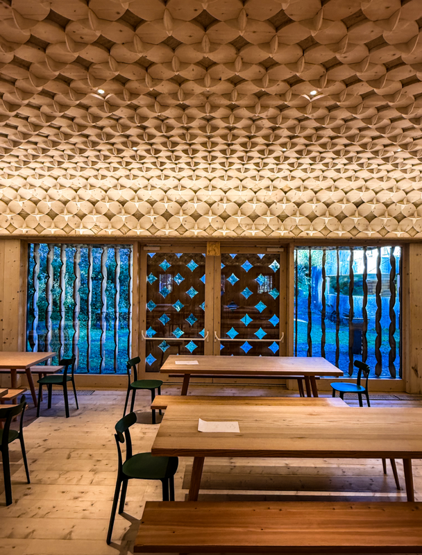 Das Bild zeigt einen großzügigen Speiseraum mit Holztischen und -stühlen, großen Fenstern und einer einzigartigen Deckenkonstruktion aus Holzpaneelen. Der Raum scheint für ein Restaurant oder eine ähnliche Gaststätte konzipiert zu sein, wobei der Schwerpunkt darauf liegt, den Gästen eine gemütliche und einladende Atmosphäre zu bieten.