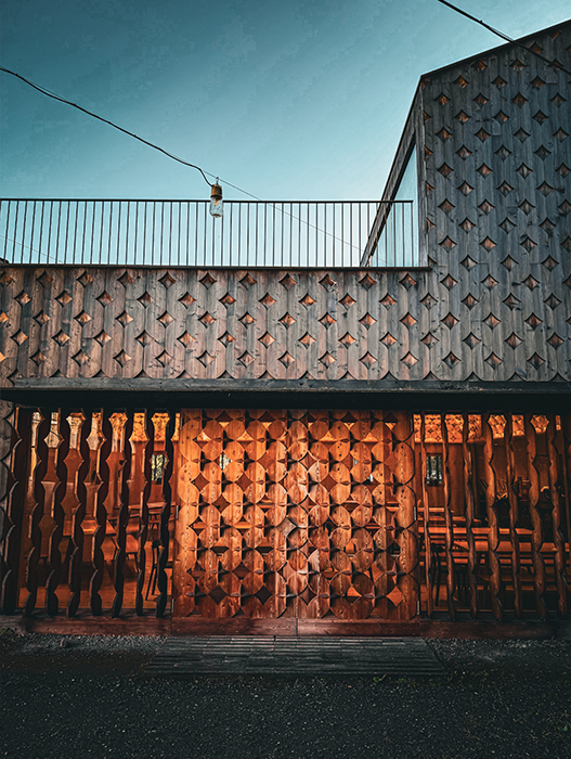 Das Bild zeigt ein Gebäude mit einer Holzfassade, die mit einem geometrischen Muster verziert ist, was ihm ein einzigartiges und ansprechendes Aussehen verleiht. Das Gebäude steht an einer Straße, auf deren linker Seite ein Geländer zu sehen ist. Die Szene wurde am Abend aufgenommen; der Himmel darüber zeigt eine Mischung aus Blau- und Orangetönen, was eine warme und angenehme Atmosphäre vermittelt.