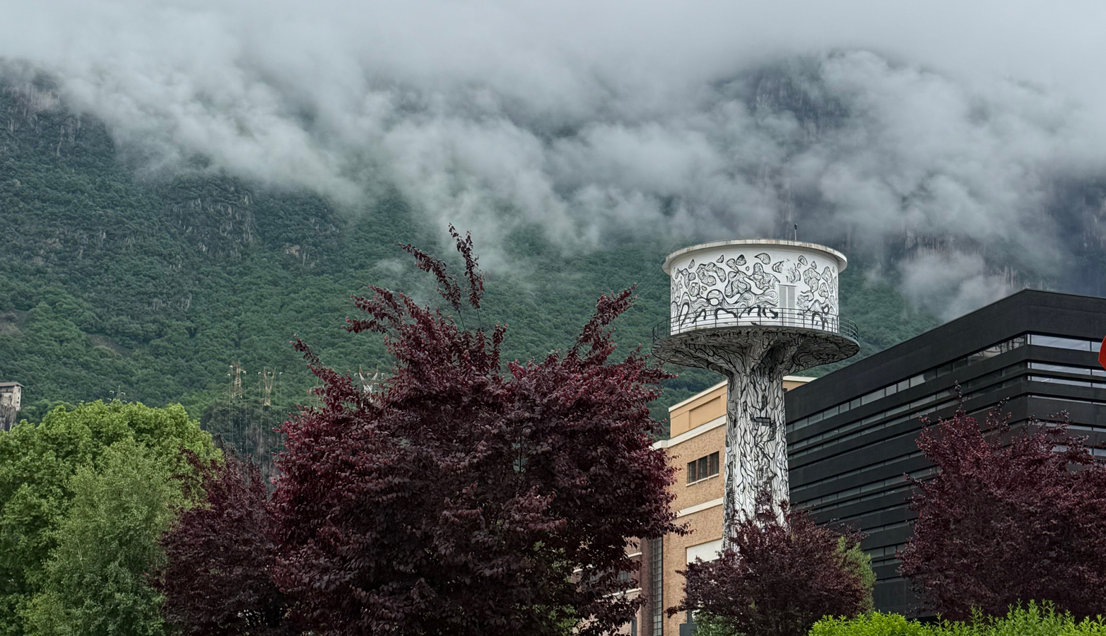 Das Bild zeigt einen Wasserturm in Schwarz-Weiß, der sich deutlich vor dem Hintergrund einer nebelverhangenen Berglandschaft im Vordergrund abhebt. Der Turm ist von üppig grünen Bäumen und Sträuchern umgeben, was eine friedliche und malerische Szenerie schafft. Die neblige Atmosphäre verleiht der Gesamtkomposition einen Hauch von Geheimnis und Ruhe.