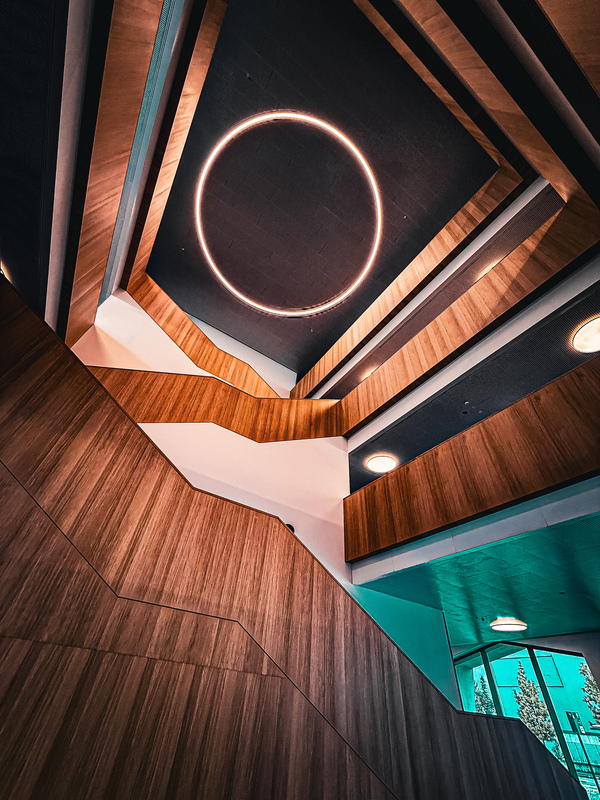 Das Bild zeigt einen Innenraum mit einer auffälligen architektonischen Gestaltung, geprägt von Holzwänden und einer runden Leuchte, die von der Decke hängt. Der Raum wirkt wie ein moderner, offen gestalteter Bereich, in dem sich natürliches und künstliches Licht vermischen. Die Perspektive des Bildes ist vom Boden aus nach oben zur Decke gerichtet, wodurch die Höhe und die Erhabenheit des Raums betont werden.