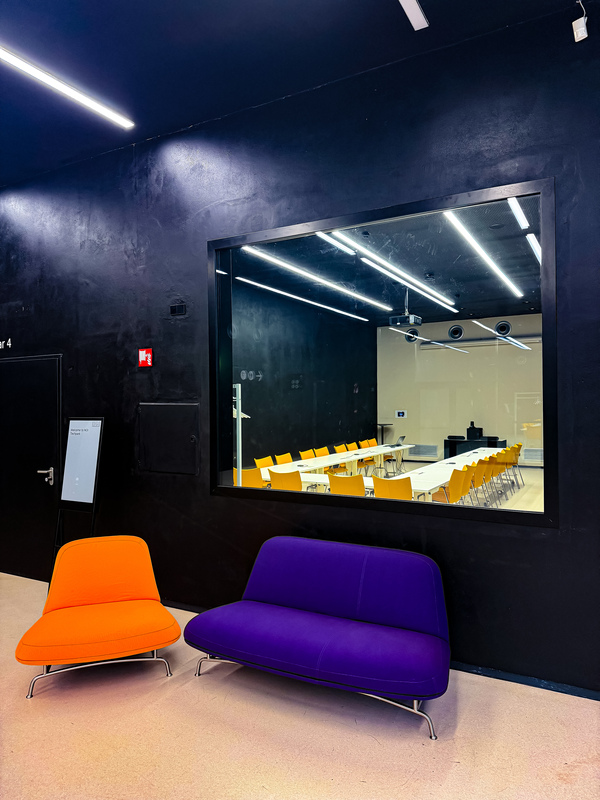 Das Bild zeigt einen modernen Büroraum mit einem großen Fenster, in dem sich ein Konferenzraum spiegelt. Das Büro verfügt über eine schwarze Wand mit einem Whiteboard und einer Tür auf der linken Seite. Im Vordergrund stehen zwei Stühle – einer in Orange und einer in Lila. Durch das Fenster ist der Konferenzraum zu sehen, in dem ein langer Tisch und Stühle für Besprechungen aufgestellt sind.