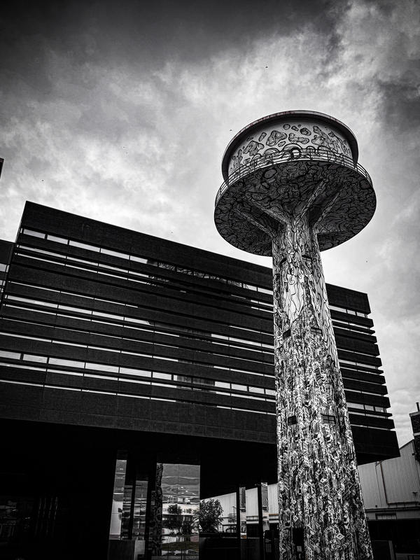 Das Bild ist eine Schwarz-Weiß-Fotografie, die einen hohen, kunstvoll verzierten Wasserturm mit rundem Dach zeigt, der vor einem modernen Gebäude mit einer gitterartigen Fassade steht. Die Aufnahme wurde aus einem niedrigen Blickwinkel gemacht, wodurch die Höhe und Erhabenheit des Turms besonders hervorgehoben werden. Die Szene scheint in einer städtischen Umgebung zu spielen, wobei das Gebäude und der Wasserturm die Hauptblickpunkte bilden.
