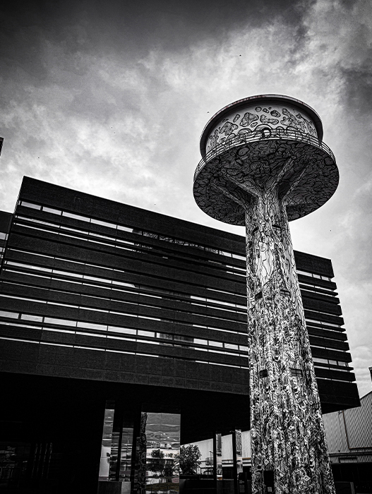 Das Bild ist eine Schwarz-Weiß-Fotografie, die einen hohen, kunstvoll verzierten Wasserturm mit rundem Dach zeigt, der vor einem modernen Gebäude mit einer Lamellenfassade steht. Die Aufnahme wurde aus einem niedrigen Blickwinkel gemacht, wodurch die Höhe und Erhabenheit des Turms betont werden. Die Fassade des Gebäudes ist im Hintergrund zu sehen und verleiht der Szene Tiefe. Das Bild enthält keine erkennbaren Texte oder zählbaren Objekte, und die relative Anordnung der Objekte lässt auf eine gut geplante Stadtlandschaft schließen.