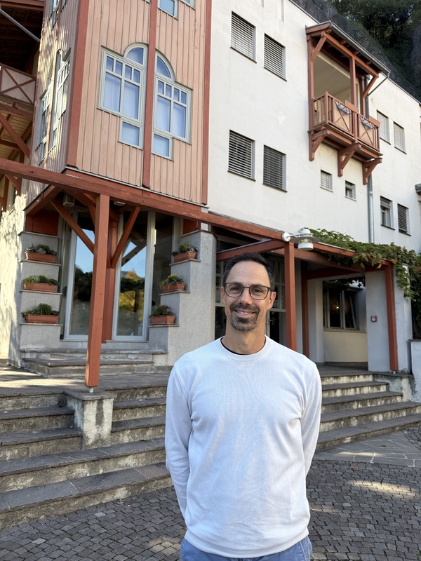 Ein Mann steht vor einem Gebäude mit Holzfassade und Balkon und lächelt in die Kamera. Das Gebäude hat mehrere Fenster und eine Tür. Der Mann trägt ein weißes Hemd und eine Brille.
