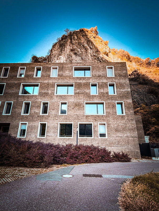 Das Bild zeigt ein modernes Gebäude mit einer einzigartigen architektonischen Gestaltung, das sich durch eine Reihe rechteckiger Fenster auszeichnet, die in einem gitterartigen Muster angeordnet sind. Das Gebäude liegt an einem Hang, im Hintergrund ist eine felsige Klippe zu sehen. Die Szene wurde aus einem niedrigen Blickwinkel aufgenommen, wodurch die Höhe des Gebäudes und seine Dominanz in der Landschaft betont werden. Das Bild ist in Farbe, mit einem strahlend blauen Himmel und etwas Grün im Vordergrund.