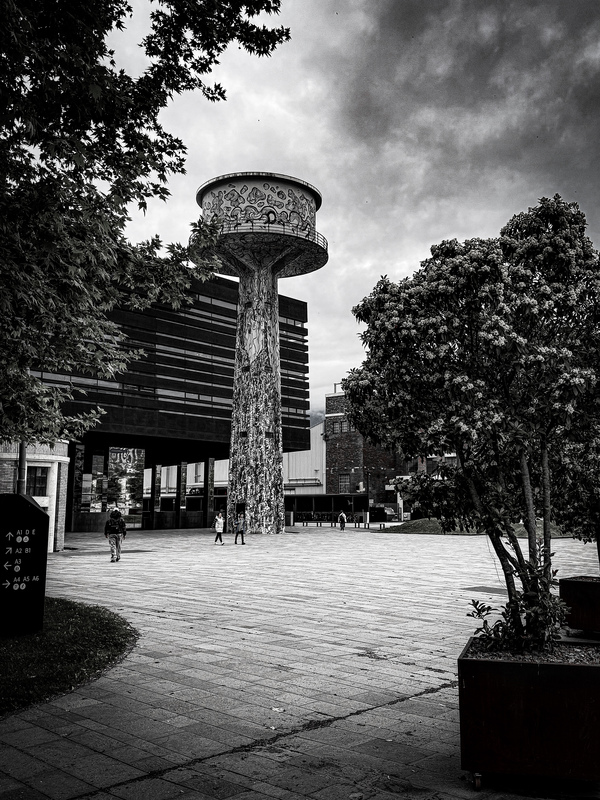 Das Bild zeigt eine Schwarz-Weiß-Fotografie eines großen, kunstvoll verzierten Wasserturms auf einem öffentlichen Platz. Der Turm ist von Bäumen umgeben, und man sieht Menschen, die in der Umgebung spazieren gehen. Die Aufnahme wurde aus einem niedrigen Blickwinkel gemacht, wodurch die Höhe und Erhabenheit des Turms besonders hervorgehoben werden. Die Gesamtkomposition und die Präsenz des Wasserturms vermitteln ein Gefühl für die Größe und Bedeutung des Bauwerks in der städtischen Landschaft.