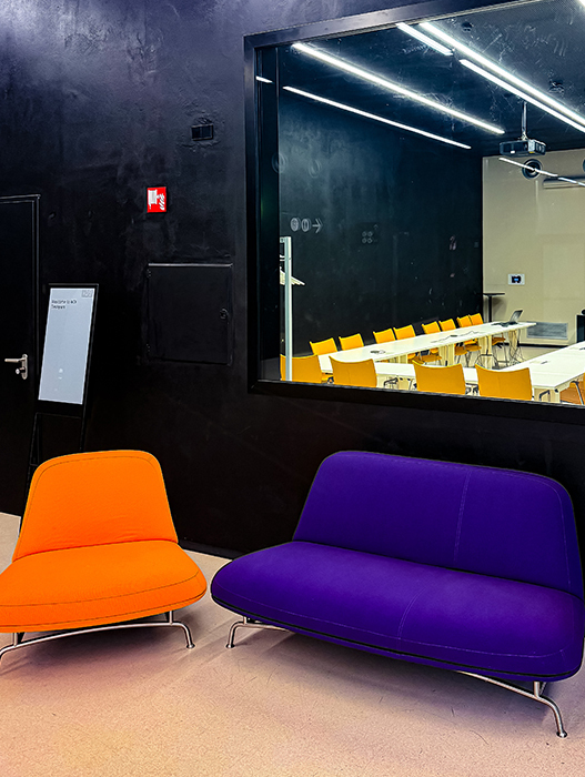 Das Bild zeigt einen modernen Büroraum mit einer schwarzen Wand und einem großen Spiegel an der Wand. Im Vordergrund stehen zwei bunte Stühle – einer in Orange und einer in Lila. Im Hintergrund ist ein Konferenzraum mit einem langen Tisch und Stühlen zu sehen, was darauf hindeutet, dass es sich um einen Raum für Besprechungen oder Präsentationen handelt.