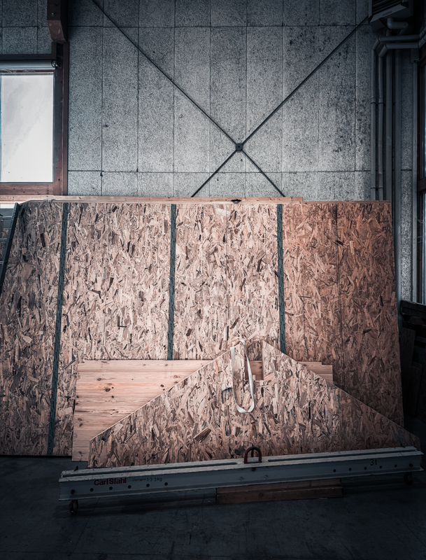 Das Bild zeigt eine Baustelle oder eine Werkstatt, in der eine große Holzplanke oder -platte auf einem Metallgestell ruht. Im Hintergrund ist eine Betonwand mit einem großen X-förmigen Muster zu sehen, und auf der linken Seite des Bildes ist ein Fenster zu erkennen. Die gesamte Szene wirkt wie eine Industrie- oder Werkstattumgebung, die möglicherweise für Bau- oder Holzbearbeitungsprojekte genutzt wird.
