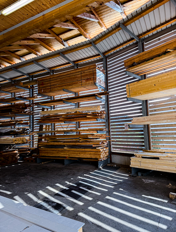 Das Bild zeigt ein großes Lagerhaus, das mit zahlreichen Holzplanken und -brettern gefüllt ist, die auf Regalen und Gestellen gestapelt sind. Das Lagerhaus verfügt über ein schräges Dach und große Fenster, durch die Tageslicht in den Raum fällt. Die Holzplanken sind in verschiedenen Stapeln und Haufen angeordnet, was einen Eindruck von Ordnung und Lagerkapazität vermittelt. Die gesamte Szene vermittelt den Eindruck eines gepflegten und funktionalen Lagerraums.