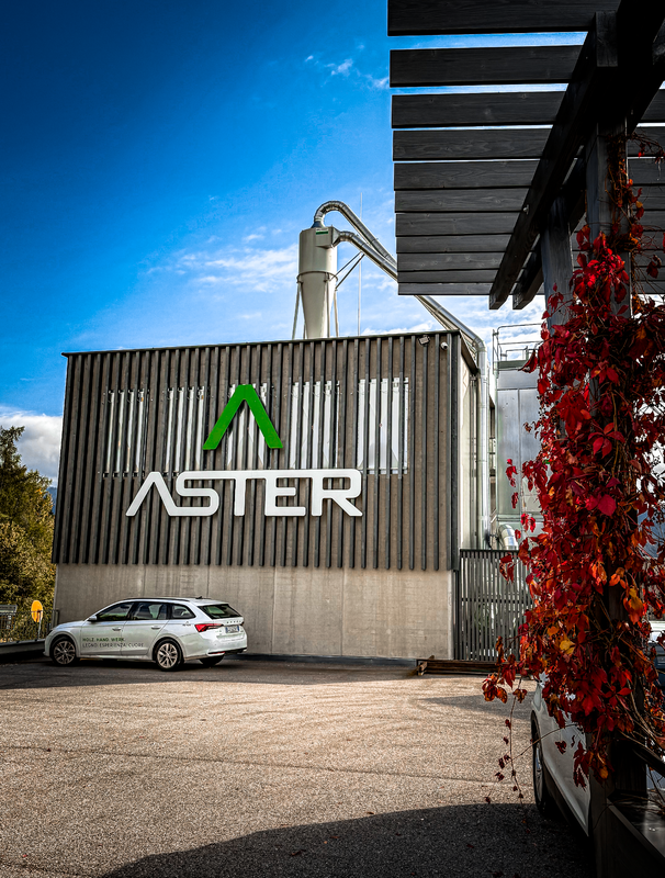 Das Bild zeigt ein großes Industriegebäude mit einem auffälligen grün-weißen Logo an der Seite, das auf das Vorhandensein eines Unternehmens oder einer Einrichtung hindeutet. Vor dem Gebäude steht ein Auto, und im Hintergrund sind einige Bäume sowie ein Holzbau zu sehen. Die Szene scheint in einer sonnigen Umgebung im Freien zu spielen.