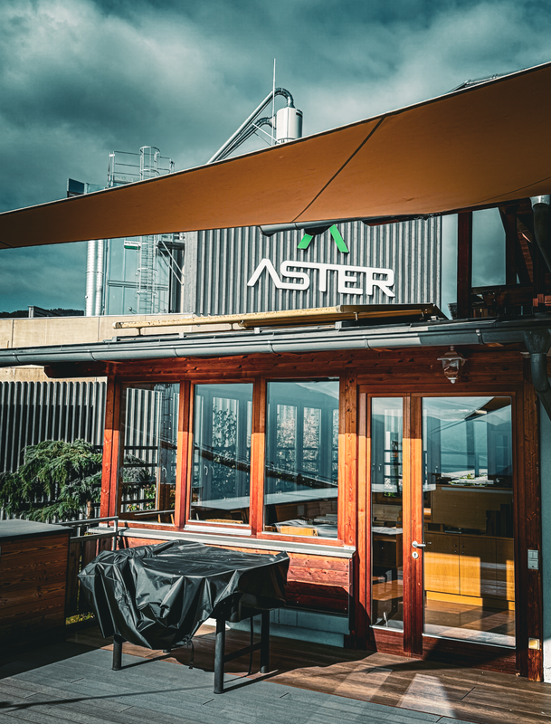Das Bild zeigt eine Holzterrasse mit einem schwarzen Tisch, der mit einer Plane abgedeckt ist, vor einem großen Gebäude mit einem auffälligen „Aster“-Schild. Die Szene spielt vor einem bewölkten Himmel, was eine etwas bedrückte Atmosphäre schafft.