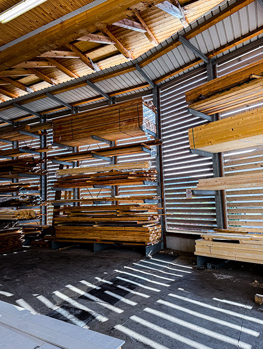Das Bild zeigt eine große Lagerhalle, die mit Stapeln aus Holzbrettern und Balken gefüllt ist, die vermutlich für Bau- oder Tischlerarbeiten verwendet werden. Die Lagerhalle verfügt über ein Wellblechdach und große Fenster, durch die Tageslicht in den Raum fällt. Die Holzbretter sind in verschiedenen Stapeln und Regalen angeordnet, was der Lagerhalle ein Gefühl von Ordnung und Struktur verleiht.