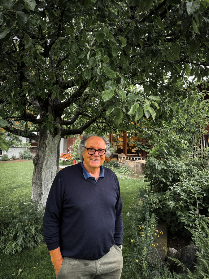Ein älterer Mann mit grauen Haaren und Brille steht in einem üppigen Garten, lächelt und blickt direkt in die Kamera. Der Garten ist voller grüner Pflanzen und Bäume, was eine ruhige und friedliche Atmosphäre schafft. Im Hintergrund ist ein Gebäude zu sehen, möglicherweise ein Haus oder ein zum Garten gehörendes Gebäude. Der Mann trägt einen blauen Pullover und eine khakifarbene Hose, die sich gut in die natürliche Umgebung einfügen.