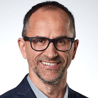Auf dem Bild ist ein lächelnder Mann zu sehen, der eine Brille und ein Sakko trägt. Er blickt mit freundlichem Gesichtsausdruck direkt in die Kamera. Der Hintergrund ist einfarbig weiß, was dazu beiträgt, die Aufmerksamkeit auf die Person zu lenken. Die Kleidung des Mannes und die Art und Weise, wie er für das Foto posiert, lassen vermuten, dass es sich um ein professionelles Porträtfoto handeln könnte.