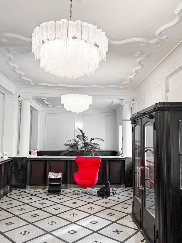 Das Bild zeigt die Lobby eines luxuriösen Hotels, in deren Mitte ein auffälliger roter Sessel steht. Der Raum verfügt über einen schwarz-weiß karierten Boden, und von der Decke hängt ein großer, mehrstufiger Kronleuchter, der den gesamten Raum in ein warmes Licht taucht. Ein schwarzer Schrank ergänzt das elegante Ambiente. Neben dem Sessel steht eine Topfpflanze, und auf dem Boden liegt ein Rucksack, was auf die Anwesenheit eines Gastes hindeutet. Das gesamte Design und die Einrichtung schaffen eine raffinierte und einladende Atmosphäre.