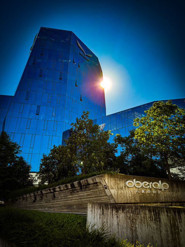 Das Bild zeigt ein modernes, bläulich schimmerndes Glasgebäude, das das Sonnenlicht reflektiert und eine visuell beeindruckende Szenerie schafft. Die Sonne scheint hell und taucht das Gebäude und die Umgebung in ein warmes Licht. Im Vordergrund steht eine Betonkonstruktion, auf der das Wort „Obelisk“ zu lesen ist, und in der Nähe ist ein Baum zu sehen. Die Gesamtkomposition des Bildes vermittelt den Eindruck einer gut durchdachten Stadtlandschaft, wobei das Glasgebäude und der Baum der Szene natürliche Elemente hinzufügen.