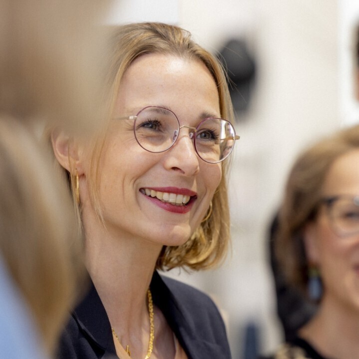Das Bild zeigt eine lächelnde Frau mit Brille und schwarzem Blazer vor einem unscharfen Hintergrund, der auf eine gesellschaftliche Veranstaltung hindeutet.