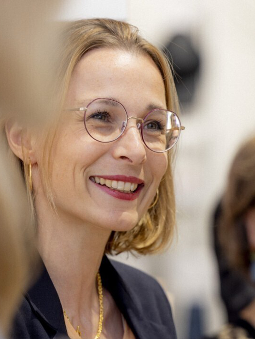 Das Bild zeigt eine Frau mit blonden Haaren, die eine Brille und einen schwarzen Blazer trägt und in die Kamera lächelt. Sie befindet sich im Vordergrund, während der Hintergrund, auf dem weitere Personen zu sehen sind, unscharf ist. Das Bild wirkt wie ein Porträt oder eine Nahaufnahme, wobei der Fokus auf dem Gesicht und dem Ausdruck der Frau liegt.