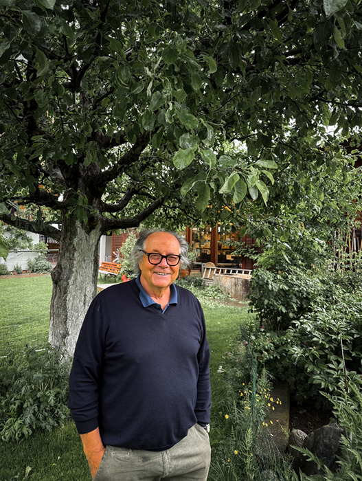 Ein älterer Mann mit Brille steht in einem üppigen Garten, lächelt und blickt direkt in die Kamera. Der Garten ist voller grüner Pflanzen, und im Hintergrund steht ein Baum.
