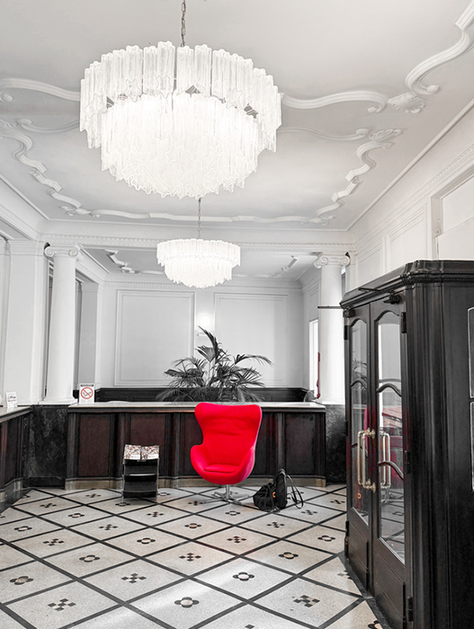 Das Bild zeigt die Lobby eines luxuriösen Hotels, in deren Mitte ein auffälliger roter Sessel steht. Der Raum verfügt über einen schwarz-weiß gefliesten Boden, und von der Decke hängt ein großer, mehrstufiger Kronleuchter, der den gesamten Raum in ein warmes Licht taucht. Auf der rechten Seite des Raumes ist ein schwarzer Schrank zu sehen, und eine Topfpflanze verleiht der Szene einen Hauch von Grün. Das gesamte Design und die Einrichtung schaffen eine elegante und einladende Atmosphäre.