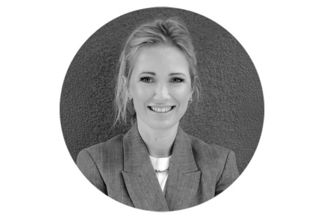 Das Bild zeigt ein Schwarz-Weiß-Porträt einer Frau mit blonden Haaren, die in die Kamera lächelt. Sie trägt einen Blazer, und man sieht eine Halskette. Der Hintergrund ist eine strukturierte Wand, die eine neutrale Kulisse für die Person bildet. Das Bild ist quadratisch gerahmt, was ihm ein klassisches und zeitloses Aussehen verleiht.