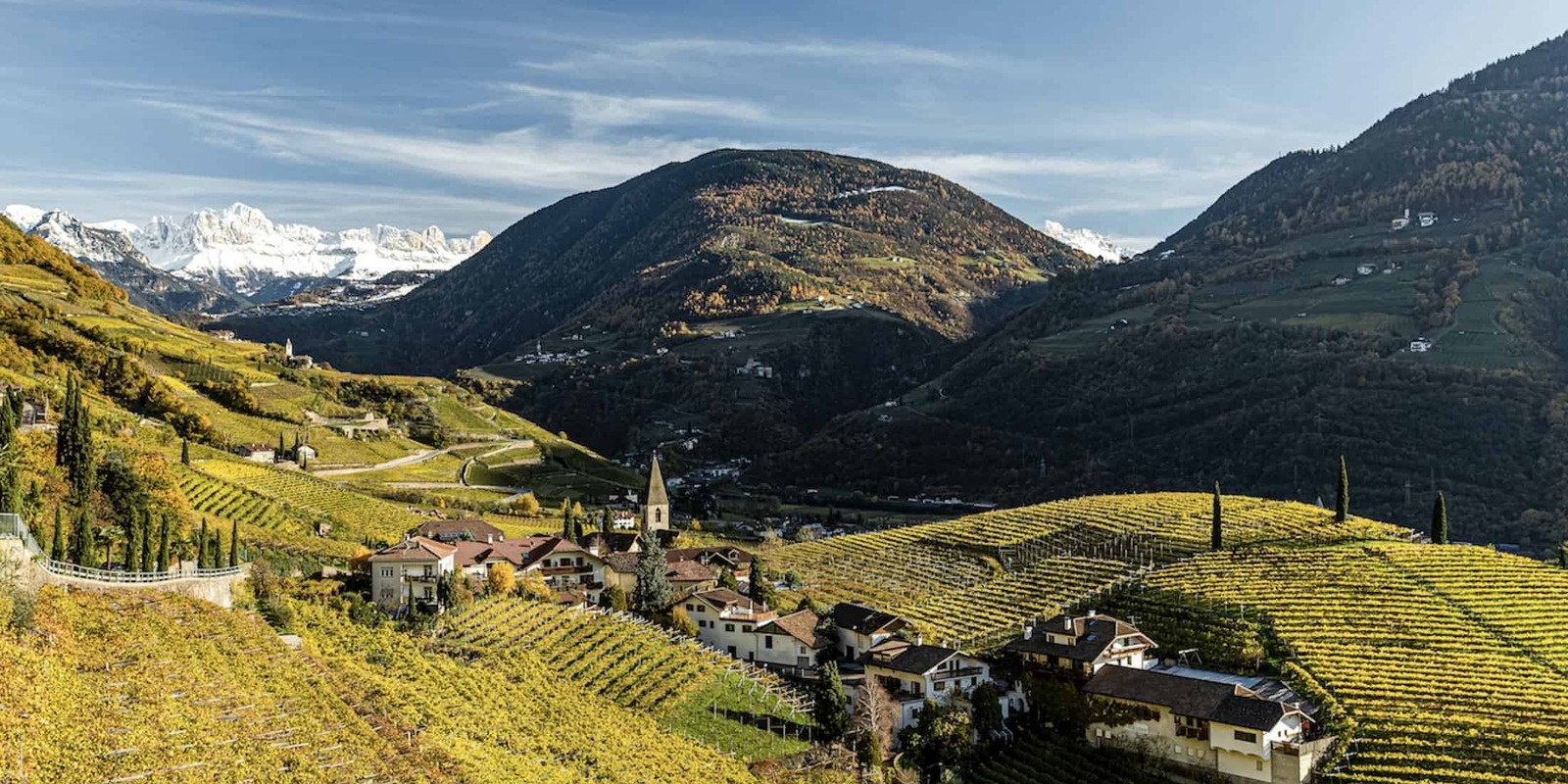 Das Bild zeigt ein malerisches Dorf, das in einem Tal liegt und von sanften Hügeln und Weinbergen umgeben ist. Das Dorf zeichnet sich durch eine Mischung aus traditioneller und moderner Architektur aus, wobei die Gebäude über die Landschaft verstreut liegen. Die Hügel sind mit üppiger grüner Vegetation bedeckt, und der Himmel darüber ist klar und blau mit einigen vereinzelten Wolken. Die Szene wurde von einem hohen Aussichtspunkt aus aufgenommen und bietet einen Panoramablick auf das Dorf und seine natürliche Umgebung.