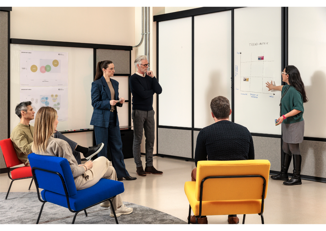 Das Bild zeigt eine Gruppe von Menschen in einer modernen Büroumgebung, wobei eine Frau vorne steht und der Gruppe etwas vorstellt. Im Raum befinden sich ein Whiteboard mit einer Grafik und ein Flipchart, und die Stühle sind im Halbkreis um die Teilnehmer herum angeordnet. Die allgemeine Atmosphäre wirkt professionell und kooperativ, und die Teilnehmer verfolgen die Präsentation aufmerksam.