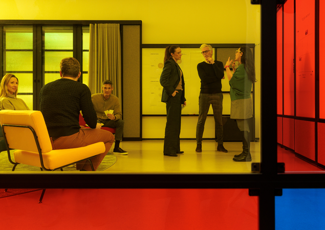 Das Bild zeigt eine Gruppe von Menschen in einem Raum mit einem gelben Stuhl, einer Weißwandtafel und einem rot-blau getönten Glasfenster. Die Personen scheinen sich in einer Diskussion oder einer Präsentation zu befinden; einige stehen, andere sitzen. Der Raum strahlt eine moderne und professionelle Atmosphäre aus, wobei die Farbkombination aus Gelb, Weiß und Rot eine optisch ansprechende Umgebung schafft.