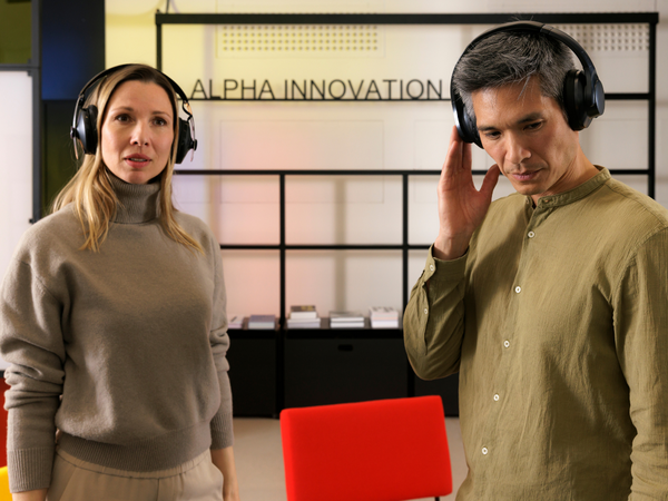 Das Bild zeigt zwei Personen mit Kopfhörern in einer Büroumgebung, im Hintergrund ist ein Schild mit der Aufschrift „ALPHA INNOVATION“ zu sehen.