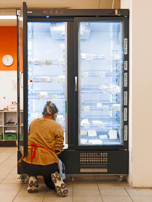 Eine Person steht vor einem großen Kühlschrank, schaut hinein und begutachtet den Inhalt. Der Kühlschrank ist mit verschiedenen Lebensmitteln gefüllt, und die Person scheint die Etiketten oder den Inhalt der Lebensmittel zu überprüfen. Die Szene spielt sich in einem Raum ab, in dem eine Uhr an der Wand hängt und in dessen Nähe sich ein Regal befindet.