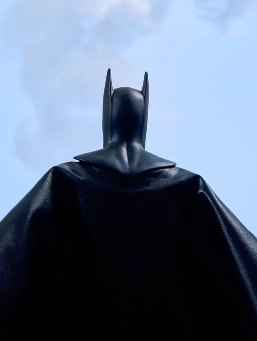 Das Bild zeigt eine schwarze Batman-Figur, die vor einem blauen Himmel mit Wolken steht. Die Figur ist mit dem Rücken zur Kamera positioniert, sodass man ihren Rücken deutlich sehen kann. Die Figur scheint aus einem glänzenden Material, möglicherweise Leder, gefertigt zu sein und steht im Mittelpunkt des Bildes.