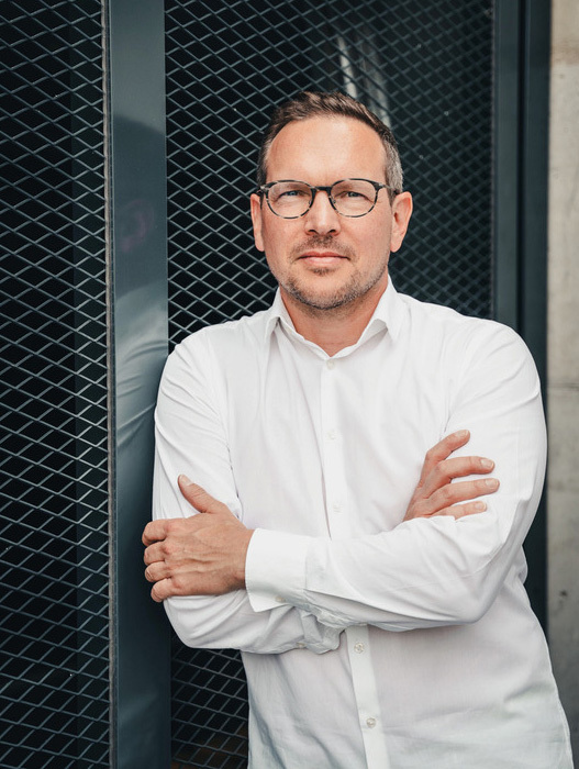 Das Bild zeigt einen Mann, der mit verschränkten Armen vor einem schwarzen Metallzaun steht. Er trägt ein weißes Hemd und eine Brille, was ihm ein professionelles Aussehen verleiht. Der Mann blickt zur Seite, möglicherweise beobachtet er etwas oder jemanden in der Ferne. Das Bild ist in Schwarz-Weiß gehalten, was der Szene einen zeitlosen Charakter verleiht.