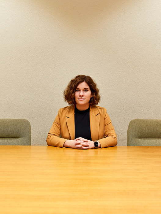 Das Bild zeigt eine Frau, die an einem Konferenztisch sitzt, einen braunen Blazer trägt und die Hände vor sich gefaltet hat. Der Tisch steht vor zwei grünen Stühlen, im Hintergrund ist eine schlichte weiße Wand zu sehen. Die Frau scheint sich in einem beruflichen Umfeld zu befinden, möglicherweise bei einer Besprechung oder einer Präsentation.