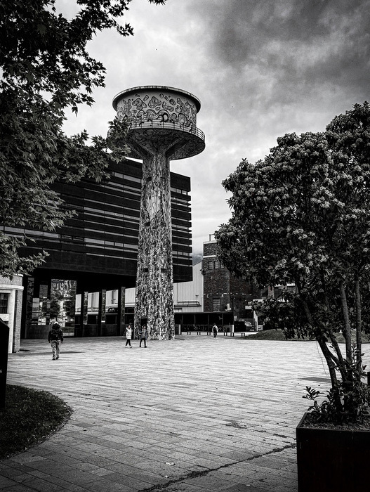 Das Bild zeigt eine Schwarz-Weiß-Fotografie eines hohen, kunstvoll verzierten Wasserturms auf einem Platz. Der Turm hat eine runde Spitze und weist ein großes, aufwendiges Design auf. Auf dem Platz sind mehrere Menschen zu sehen, die umhergehen, und im Hintergrund sind Bäume und ein Gebäude zu erkennen. Die Aufnahme wurde aus einem niedrigen Blickwinkel gemacht, wodurch die Höhe und Erhabenheit des Turms besonders hervorgehoben werden.