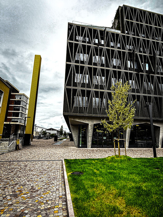 Das Bild zeigt ein modernes Gebäude mit einer geometrischen Fassade, die sich durch eine Mischung aus Glas- und Betonelementen auszeichnet. Das Gebäude steht auf einem gepflasterten Bereich, im Vordergrund befindet sich eine Rasenfläche. Ein Baum ist in der Szene zu sehen und verleiht der städtischen Umgebung einen Hauch von Natur. Der Himmel darüber ist bewölkt, was eine stimmungsvolle Atmosphäre schafft. Das Bild wurde aus einem niedrigen Blickwinkel aufgenommen, wodurch die Höhe und die architektonische Gestaltung des Gebäudes betont werden.